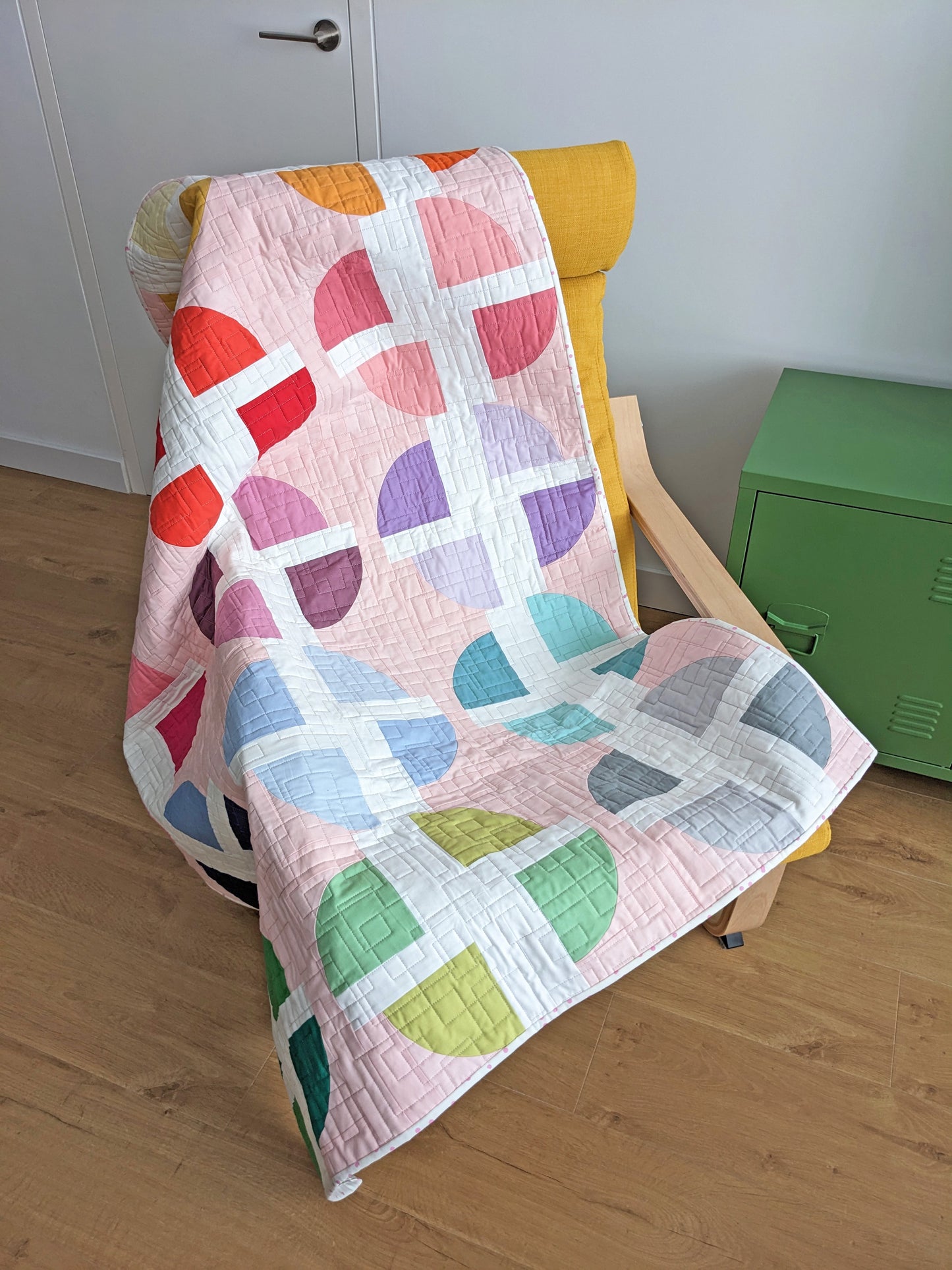 A pastel geometric quilt draped over a yellow armchair in a room with light wood floors, a white door, and a green storage cabinet.