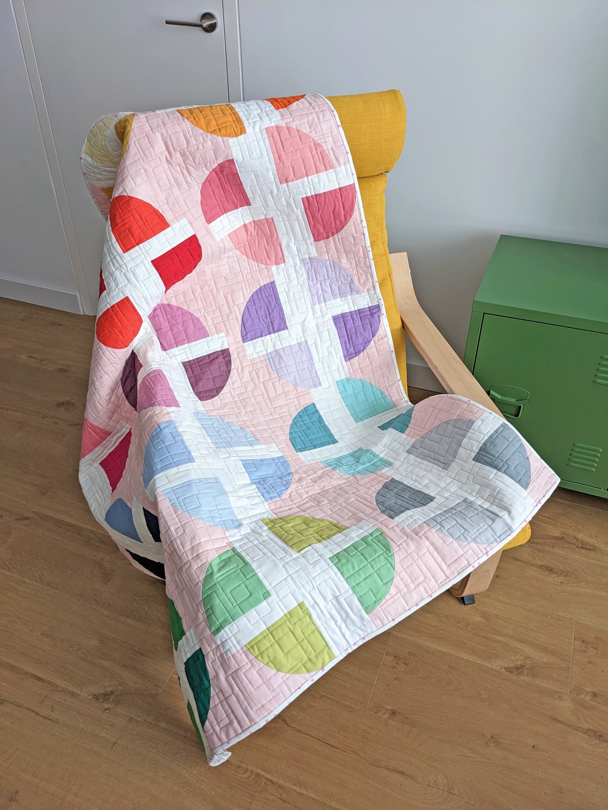 A pastel geometric quilt draped over a yellow armchair in a room with light wood floors, a white door, and a green storage cabinet.
